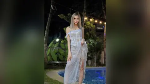 Elegant blonde woman in a white sheer lace gown standing by a tropical poolside at night.