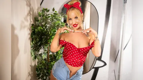 Smiling blonde woman in a red polka dot top and denim shorts posing in a bright modern room.