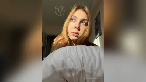Blonde woman in cozy loungewear looking thoughtful in warm, sunlit bedroom lighting.