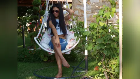 Smiling woman in a white tank top and denim shorts relaxing in a hanging garden chair on a sunny day.