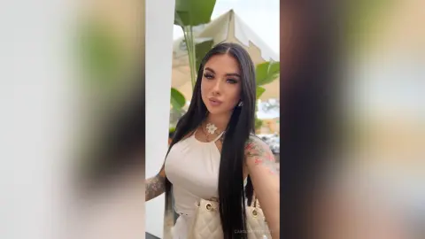 Young woman with long dark hair wearing a white sleeveless top and necklace in a bright outdoor setting.