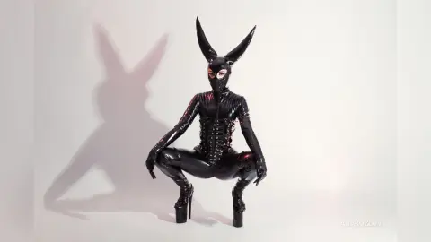 Woman in black latex bunny suit and high heels posing in a minimalist studio setting.