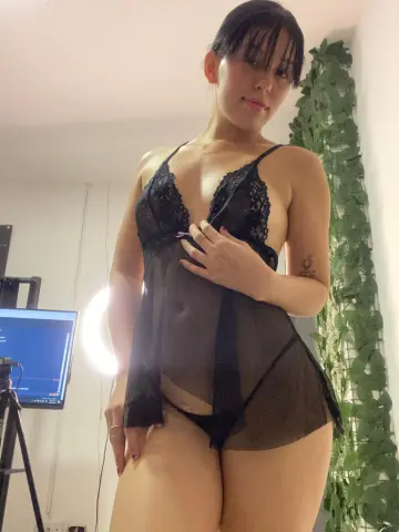 Young woman in black lace lingerie posing in a bright room with green leafy decor.