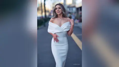 Elegant woman in a white off-the-shoulder evening gown posing on a city street at sunset.
