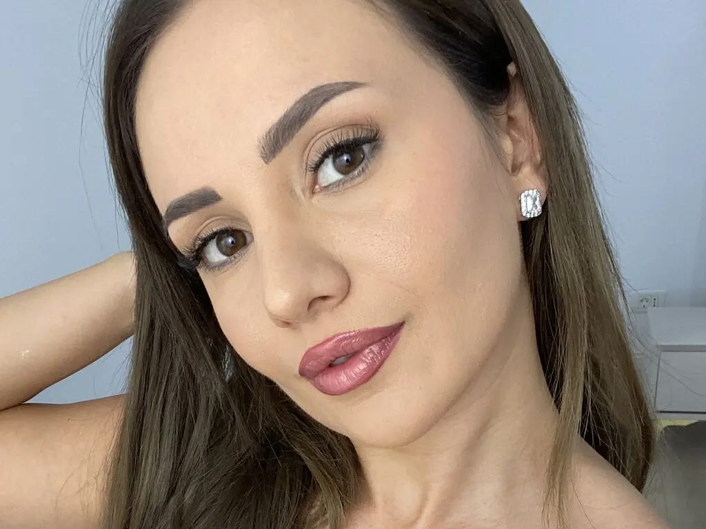 DeviaTelerr poses close-up with long brown hair, wearing silver stud earrings and pink lipstick against a plain light background.