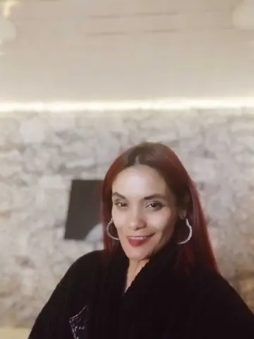 Smiling woman with red hair wearing a black sweater in a cozy room with a stone wall background.