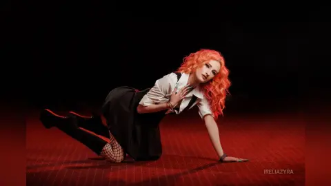 Woman with wavy orange hair in a white shirt and black skirt posing against a dark red background.