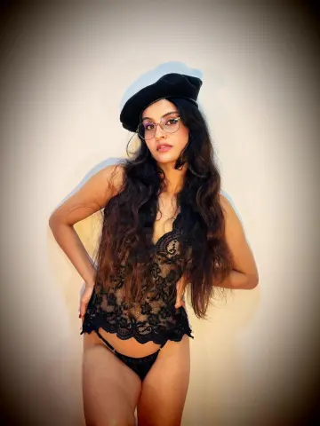 Young woman with long wavy hair wearing a black lace bodysuit and beret posing against a plain wall.