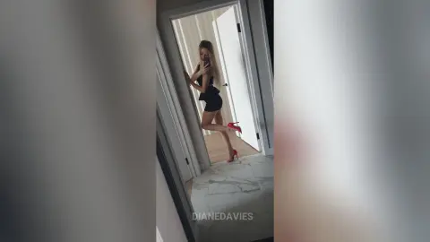 DianeDavies poses in a black outfit and red high heels, standing in a doorway against a white wall.