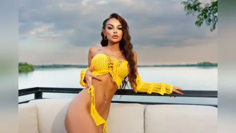 Anastasia poses in a yellow crochet bikini top and bottomset against a lake background.
