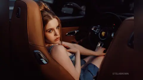 StelaBrown sits in a car seat wearing a denim top, looking back over her shoulder with her hair tied up.
