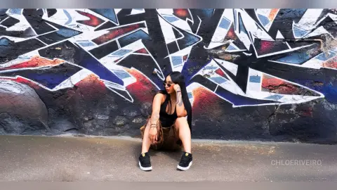 ChloeRiveiro — Stylish woman in a black tank top and brown shorts sitting against a vibrant urban graffiti wall.