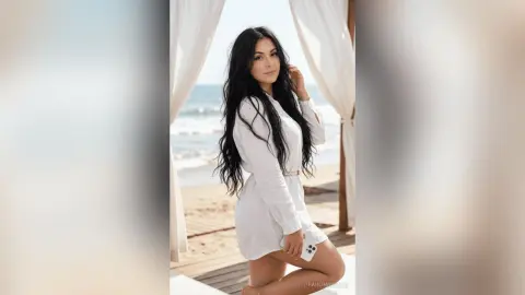 Smiling woman in a white linen dress relaxing on a sunny beach balcony overlooking the ocean.