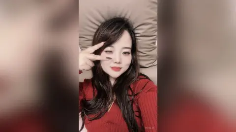 EnyaLin poses with a peace sign, wearing a red long-sleeve top while leaning against a light brown pillow.