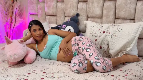 Smiling young woman in light blue tank top and pink penguin pajamas relaxing on a bed with plush toys.