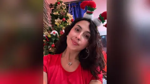 RossiNoleen smiles wearing a red t-shirt and a red and white elf hat next to a decorated Christmas tree.