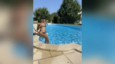 Young woman in a patterned bikini relaxing by a bright blue swimming pool on a sunny summer day.