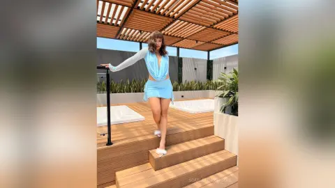 Young woman in a light blue mini dress posing on a wooden deck by a luxury rooftop pool under a pergola.