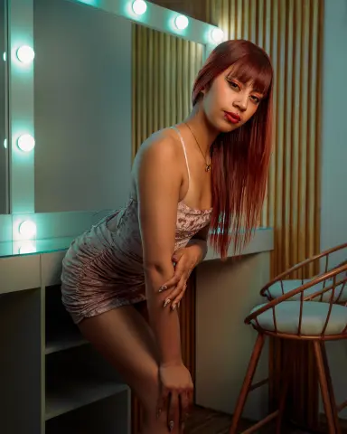 Woman in a pink floral mini dress posing in a lit vanity room with a warm, moody atmosphere.