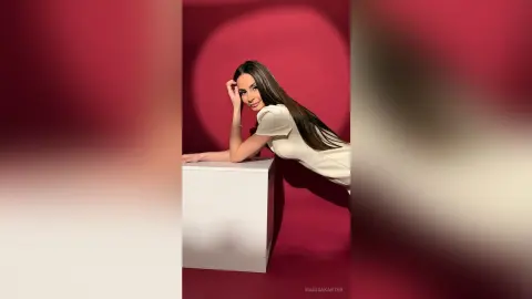 MarisaKarter poses with long straight hair, wearing a white short-sleeved top while leaning against a white block against a red background.