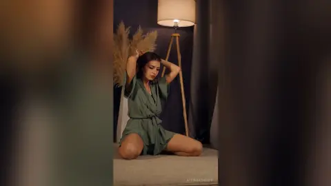 Young woman in a green satin loungewear set posing in a moody, dimly lit room with pampas grass decor.