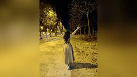 Woman in a beige midi dress standing with arms raised on a lit park path at night.
