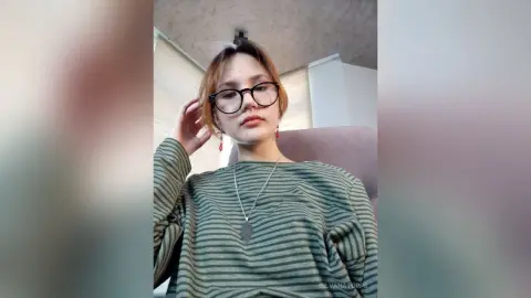 Young woman with glasses and striped green sweater posing in a bright indoor setting.