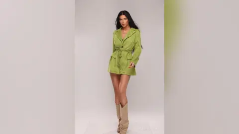 Elegant woman in a lime green belted satin mini dress and beige boots posing against a neutral studio background.