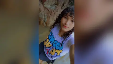 Young woman with curly hair in a graphic white t-shirt relaxing by a rocky natural spring.