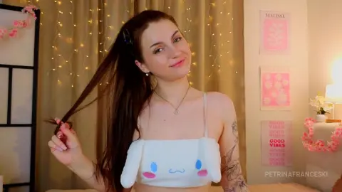 PetrinaFranceski smiles while holding a strand of her long brown hair, wearing a white bunny-eared tank top in a room with warm fairy lights.
