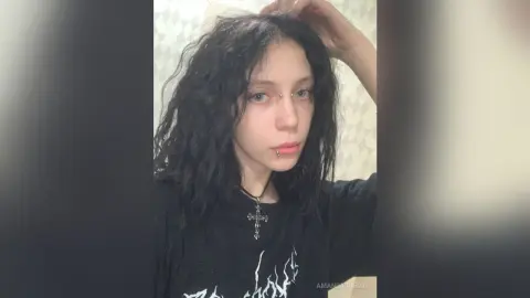 Young woman with wavy dark hair wearing a black graphic t-shirt and silver cross necklace in a soft indoor setting.