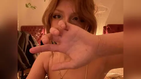 Blonde woman in a gold top making a hand gesture in a cozy, dimly lit bedroom setting.