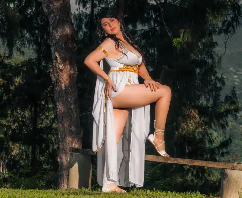 Elegant woman in a white and gold Greek goddess gown posing gracefully in a lush forest setting.