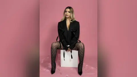 Elegant woman in a black blazer and lace tights sitting against a pink background with a chic, stylish mood.