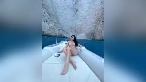 Woman in a bikini relaxing on a white boat deck against a backdrop of dramatic coastal cliffs and blue sea.