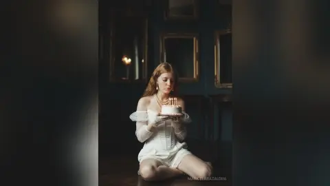Elegant woman in white satin outfit holding a small cake in a moody, vintage dark academia room.