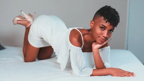 AnnPrice poses on a white bed wearing a white off-the-shoulder top and skirt, looking at the camera with short dark hair and gold hoop earrings.