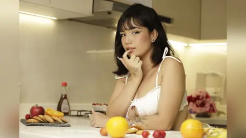Young woman in a white summer top sitting in a bright kitchen with fruit and snacks on the counter.