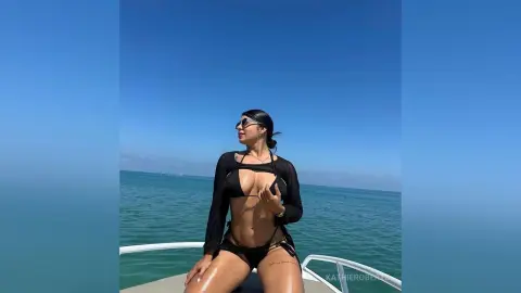 Woman in black bikini and sheer cover-up relaxing on a boat under a clear blue sky by the ocean.