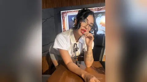 Stylish woman in a white button-down shirt and glasses posing confidently at a wooden desk.