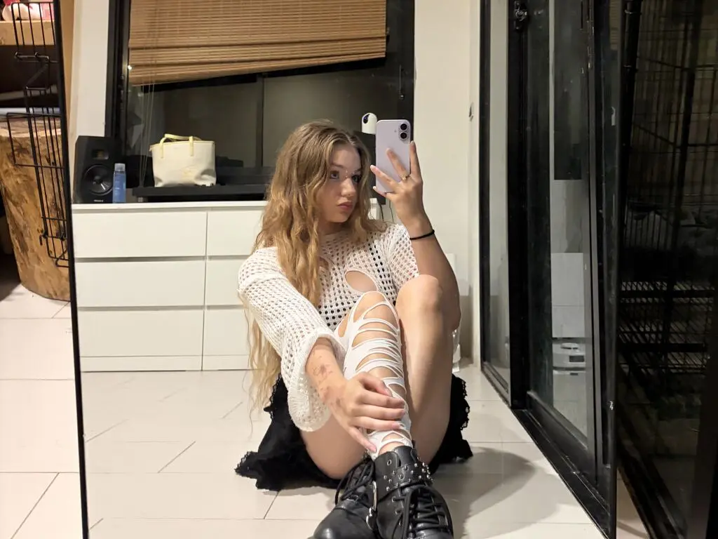 Young woman in white knit sweater and black skirt sitting on floor taking a mirror selfie in a modern room.