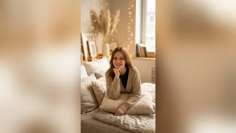 Smiling young woman in a beige cardigan relaxing on a cozy bed in a warm, fairy-lit bedroom setting.