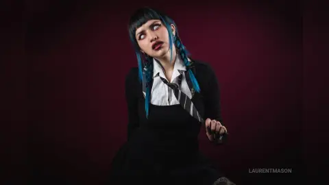 Gothic woman with blue braided hair in a black dress and tie posing against a dark moody background.