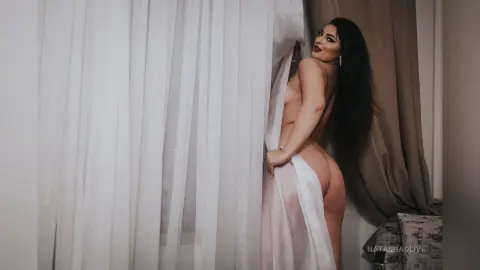 Sensual woman in a white sheer robe posing by curtains in a soft, intimate indoor setting.