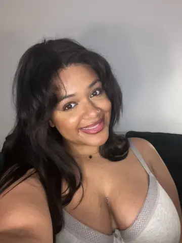 Hazelvalentine smiles, showing her long dark hair and cleavage while wearing a white lace bra against a plain wall.