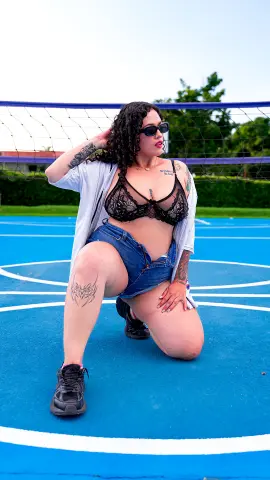 Curvy woman with tattoos in a black lace bra and denim shorts posing on a blue outdoor sports court.