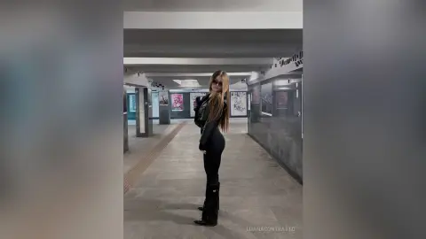 LuanaContratto stands in a hallway wearing a black long-sleeved top, black leggings, and black boots with her long hair down.