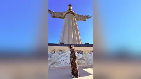 Woman in a long leopard print dress standing before the Christ the Redeemer statue under a clear blue sky.