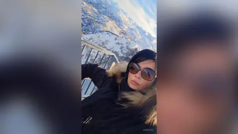 Woman in a black winter jacket and sunglasses relaxing against a snowy mountain landscape on a sunny day.