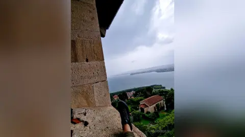 Person in dark clothing overlooking a scenic coastal town and sea from a rustic stone balcony under a cloudy sky.